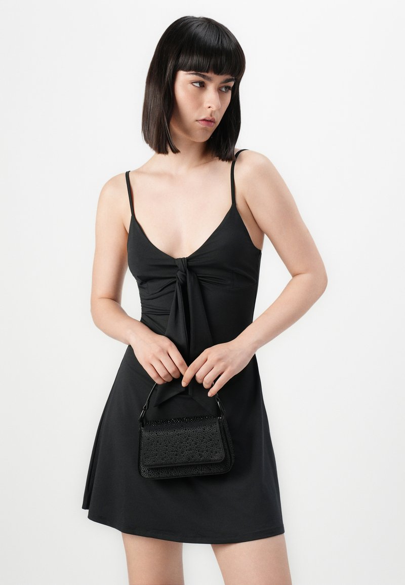 Abito nero con un dettaglio a fiocco sul décolleté, realizzato in tessuto morbido e con una gonna svasata. Accompagnato da una piccola borsa nera dal tessuto strutturato.