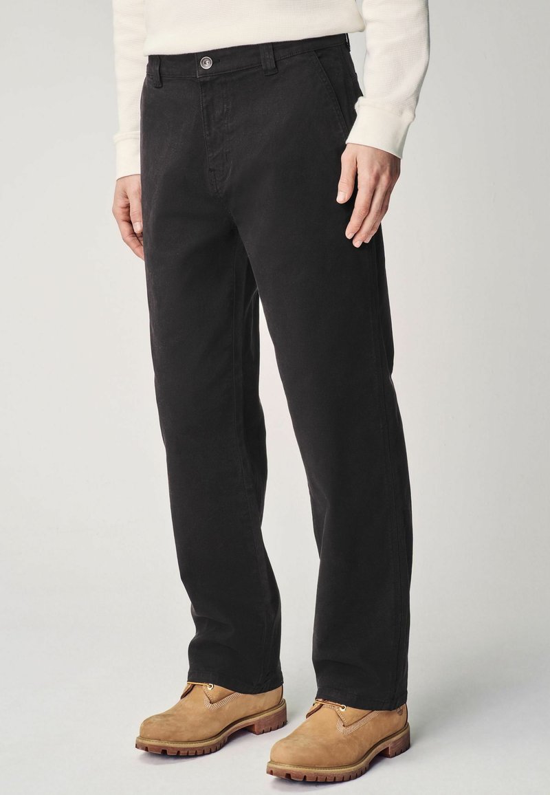 Zwarte corduroy broek met een recht model, voorzien van een voorste knoopsluiting en zijzakken. Draagt een paar beige laarzen.