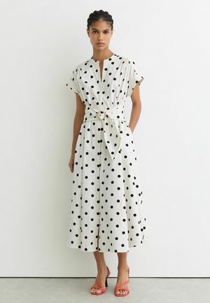 Femme portant une robe midi blanche à pois noirs, manches courtes, ceinture à la taille, et sandales corail à talons hauts, debout contre un mur blanc.