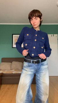 Tamsiai mėlynas megztas kardiganas su auksiniais mygtukais ir dviem priekinėmis kišenėmis, derinamas su šviesiai mėlynais plačiomis kojomis džinsais, kuriuose yra blukęs raštas.