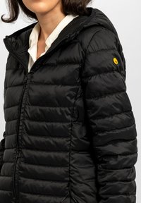 Giacca piumino nera con cappuccio, con design trapuntato, chiusura con zip e piccolo logo giallo sulla spalla sinistra. Texture liscia.