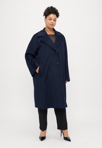 Cappotto blu navy con davanti doppiopetto, ampi revers e tasche laterali. Il tessuto strutturato conferisce un aspetto rigoroso. Indossato sopra pantaloni neri.