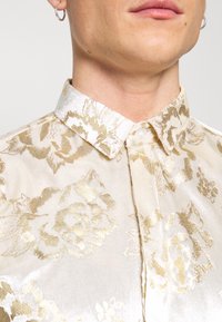 Camicia in tessuto setoso con motivo floreale dorato, dotata di colletto bianco, chiusura con bottoni e una lieve lucentezza. Vista ravvicinata del collo e della trama del tessuto.