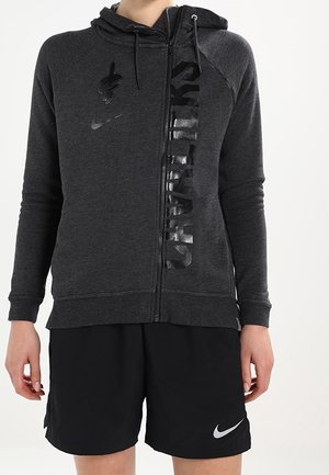 Osoba nosząca ciemnoszarą bluzę z kapturem na zamek błyskawiczny z czarnym pionowym napisem oraz czarne spodenki Nike z białym logo typu swoosh.