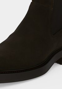 Gros plan d’une botte Chelsea en daim brun foncé montrant la tige lisse, le panneau latéral élastique et le petit talon superposé.