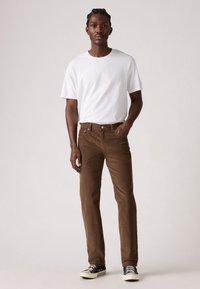 Pantaloni in corduroy marroni con un fit dritto, abbinati a una maglietta a maniche corte bianca e a scarpe sneakers nere basse. Pose in piedi, sfondo neutro.