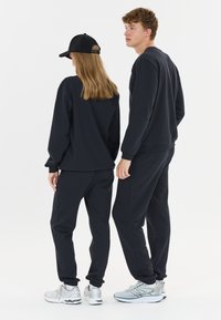 Zwarte katoenen sweatshirt en joggers met een relaxte pasvorm. De joggers hebben elastische boorden. Een zwart pet wordt gedragen door één persoon.