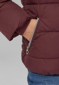 Bordo puffer jakna s patentnim zatvaračem, s teksturiranom tkaninom i podstavljenim dizajnom; traperice su vidljive ispod.