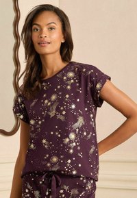 Camiseta de pijama de manga corta en color púrpura con estampado de estrellas doradas y cometas; cuenta con un cuello redondo y un corte relajado con puños enrollados.