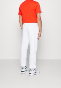 Červené atletické tričko s krátkým rukávem a postranními pruhy, v kombinaci s bílými tepláky. Boty jsou bílé se zelenými detaily a logem na patě.