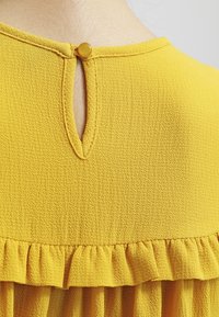 Top giallo con tessuto testurizzato, orlo arricciato, apertura a orecchino sul retro e chiusura con bottone rotondo.