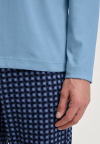 Chemise bleu clair à manches longues avec une texture douce, accompagnée d'un pantalon à motifs sombres présentant des carrés bleus et violets sur un fond marine.