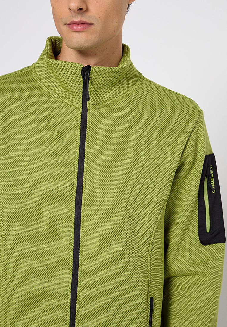 Grüne Zip-Jacke mit strukturiertem Muster, schwarzem Reißverschluss und einer Reißverschlusstasche am linken Ärmel mit einem grünen Akzent.