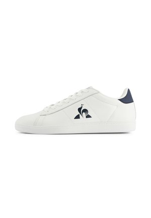Baskets basses blanches avec des lacets blancs, un empiècement au talon bleu marine et un logo de coq noir sur le panneau latéral.