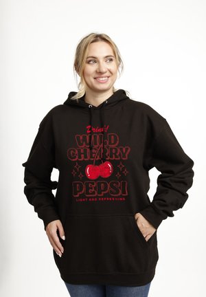 Femme souriante portant un hoodie noir avec le logo rouge "Wild Cherry Pepsi" et deux cerises, les mains dans la poche avant, fond clair.