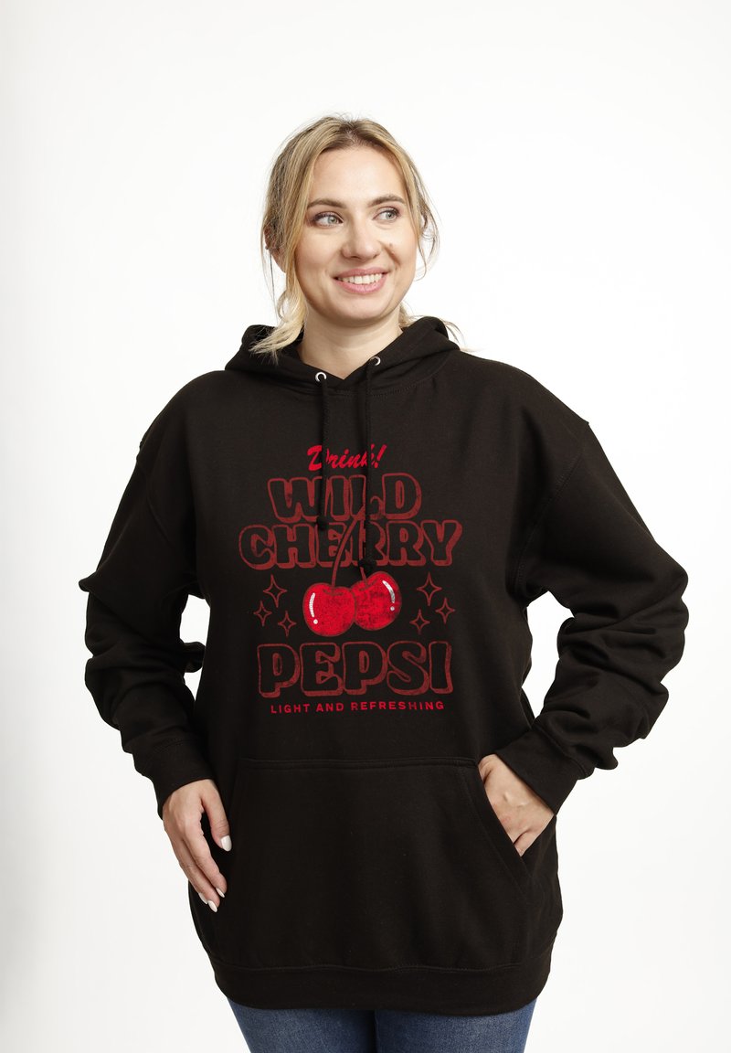 Donna che sorride indossando una felpa nera con il logo rosso "Wild Cherry Pepsi" e due ciliegie, mani nella tasca frontale, sfondo chiaro.