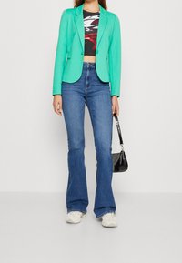 Kobieta w turkusowej marynarce, abstrakcyjnym topie crop, niebieskich dzwonach, białych sneakersach, trzymająca małą czarną torebkę na tle białym.