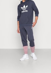 Sudadera navy con logo blanco de Adidas, combinada con pantalones de chándal navy que tienen acentos rosas y franjas laterales blancas. Textura de ante, corte relajado.