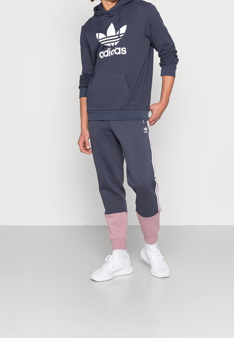 Sudadera navy con logo blanco de Adidas, combinada con pantalones de chándal navy que tienen acentos rosas y franjas laterales blancas. Textura de ante, corte relajado.