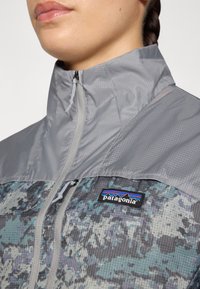Person wearing a gray and camo-patterned Patagonia jacket with a high collar and front zipper, showing the brand logo on the chest.