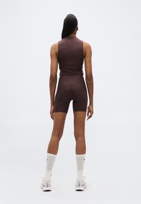 Γυναίκα που στέκεται με πλάτη, φοράει ένα αμάνικο καφέ τοπ, αντίστοιχα σορτς ποδηλασίας, λευκές κάλτσες μέσης και λευκά αθλητικά παπούτσια σε απλό φόντο.