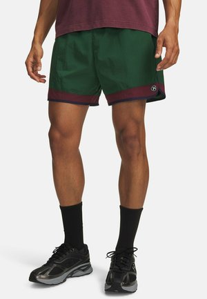 Mann trägt dunkelgrüne Sportshorts mit kastanienbraunem und schwarzem Besatz, schwarze Sportsocken und schwarze Laufschuhe und steht vor weißem Hintergrund.