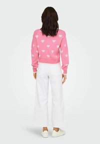 Roza pulover z belimi srčastimi vzorci, rebrastim robom in rokavi. Kombiniran z belimi širokimi hlačami in belimi supergami.