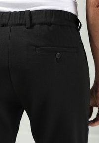 Zwarte broek met een slank ontwerp, elastische tailleband en een enkele achterzak met knoopsluiting. Soepele stoftextuur.