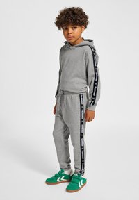 Grå huvtröja och joggers med svarta sidostråk som har en logotyp. Gröna sneakers med vita detaljer. Mjuk, avslappnad tygkvalitet och lös passform.