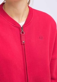 Sweat-shirt rouge à fermeture éclair, fabriqué en tissu doux, doté d'un col côtelé et d'un logo brodé sur le côté gauche de la poitrine. Texture lisse.