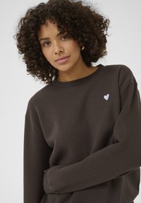 Hnědá mikina s kulatým výstřihem a oversized střihem. Obsahuje malé bílé vyšívané srdíčko na prsou. Měkká textura.