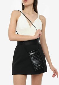 Sort faux læder crossbody taske med krokodillemønster, rektangulær form og tynd rem, holdt mod en kontrasterende hvid top og sort nederdel.