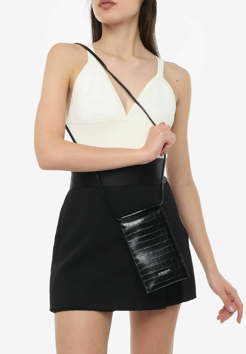 Sort faux læder crossbody taske med krokodillemønster, rektangulær form og tynd rem, holdt mod en kontrasterende hvid top og sort nederdel.