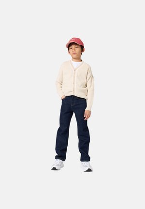 Garçon portant un cardigan beige, une chemise blanche, un jean bleu foncé, des baskets blanches et une casquette rouge, debout avec une main dans la poche.