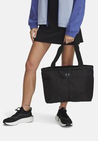 Zwarte tote bag met een gewatteerde textuur, met een voorvak en logo, vastgehouden door een persoon die zwarte schoenen en een lichte jas draagt.