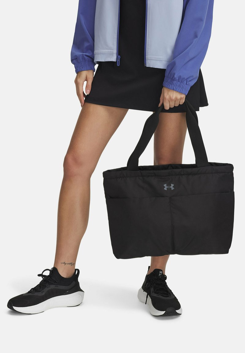 Zwarte tote bag met een gewatteerde textuur, met een voorvak en logo, vastgehouden door een persoon die zwarte schoenen en een lichte jas draagt.