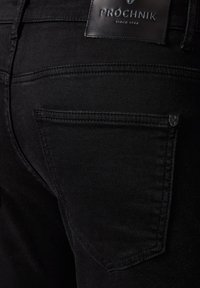 Zbliżenie na czarne dżinsy, pokazujące tylną kieszeń oraz skórzaną metkę z logo "PROCHNIK" przyszytą nad kieszenią.