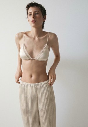 Vrouw met kort haar die een crèmekleurige bralette met kanten rand en beige geplooide broek draagt tegen een effen lichte achtergrond.