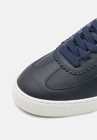 Adidași din piele navy cu partea superioară texturată, talpă din cauciuc alb contrastant și design cu șiret plat. Prezintă margini ondulate de-a lungul vârfului.