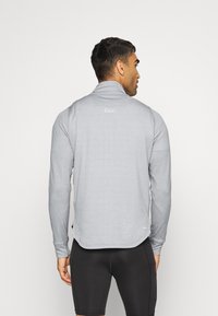 Homem de costas, vestindo uma camisola de corrida de manga longa cinza claro e calções de desporto pretos, contra um fundo branco liso.