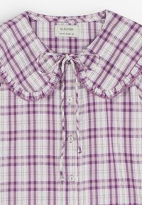 Blusa a cuadros púrpura y blanca con cuello fruncido, cierre de botones y un lazo atado en el escote. Hecha de tela ligera.