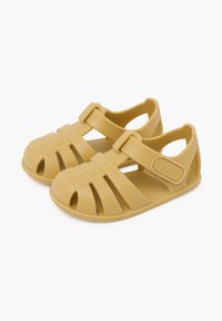 Sandalias de plástico amarillas con punta cerrada, lados abiertos y diseño de tiras cruzadas. Incluyen una correa ajustable y un acento de logo elevado. Textura suave.