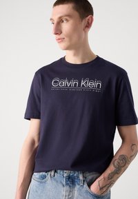 Donnarblauwe katoenen t-shirt met het "Calvin Klein" logo in het wit op de borst, ronde halslijn en korte mouwen, gecombineerd met een lichte denim jeans.