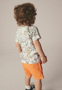 Bambino con capelli ricci che indossa una maglietta bianca con grafica e pantaloni corti arancioni, in piedi di spalle contro uno sfondo neutro.