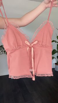 Camiseta de color durazno con adornos de volantes, lazo de cinta en el frente y tirantes finos. Tejido suave con un dobladillo delicado y transparente.
