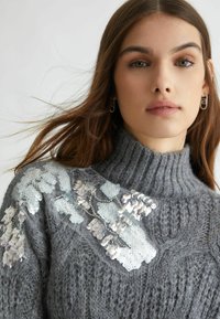 Maglione grigio lavorato a maglia con un collo alto, decorato con paillettes bianche e argentate sovrapposte che creano un motivo floreale su una spalla.