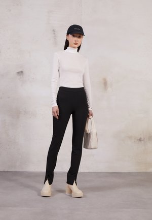 Hvid langærmet turtleneck, sorte skræddersyede bukser med slids, beige platform sko og en lys beige håndtaske. Sort kasket med logo.