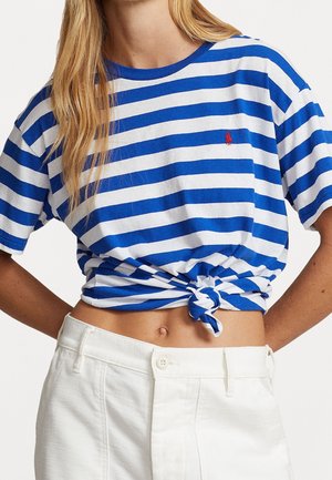 Femme portant un crop top rayé bleu et blanc noué à la taille et un pantalon blanc taille haute, bras derrière le dos.