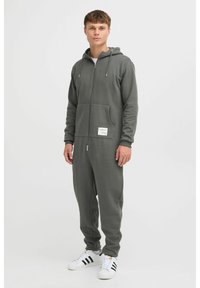 Grauer Zip-Up-Hoodie-Jumpsuit mit langen Ärmeln, Fronttasche und Kordelzugkapuze, gefertigt aus weichem Stoff. Kombiniert mit weißen Turnschuhen.
