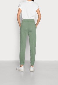 Joggers de algodón verdes con cintura elástica y bolsillos laterales, que presentan piernas ajustadas y costuras en contraste. Combinados con zapatillas blancas.